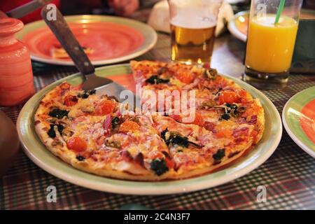 Fresh pizza with tomatoes, cheese, pepperoni, olive and mushrooms. Traditional italian pizza on dish plate with spatula in side view. Stock Photo