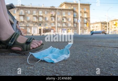 abandoned and used antibacterial medical mask lies on the asphalt at the feet of a man who is sitting on a bench in a city Park Stock Photo