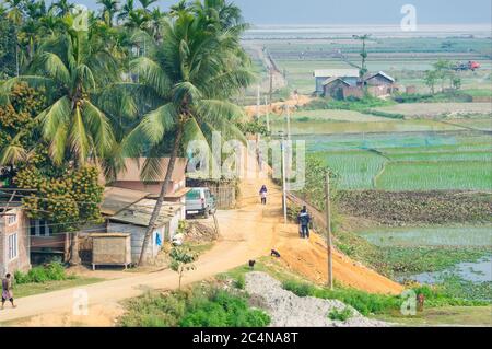 Village in Assam, India with villagers walking along the road, near rice fields Stock Photo