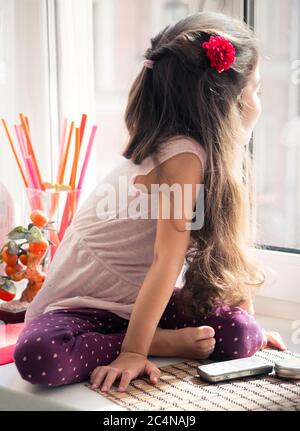 Long-haired curly little girl sitting on window-sill looking out the window. Stock Photo