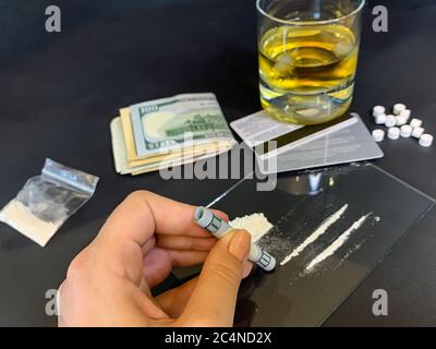 A female's hand holding rolled 100 us dollars banknote near two lines of cocaine. Plastic bag with illegal drugs, tablets, money and a glass of whiske Stock Photo