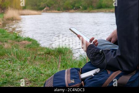 Man uses smartphone while charging from the power bank on the river's bank. Modern technology concept. Selective focus. Stock Photo