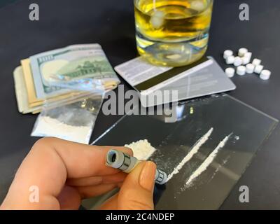 A female's hand holding rolled 100 us dollars banknote near two lines of cocaine. Plastic bag with illegal drugs, tablets, money and a glass of whiske Stock Photo