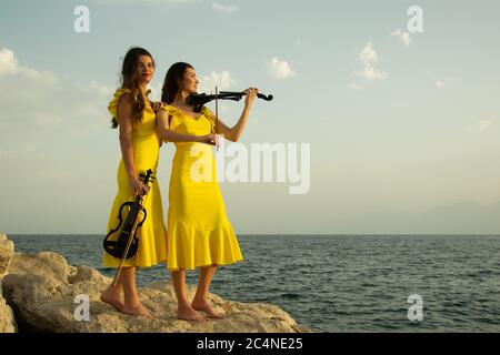 Two beautiful girls violinists in yellow concert dresses are playing electric violins with sunset, mountains and Mediterranean sea on the background. Stock Photo