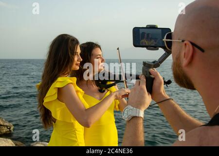 Two beautiful girls violinists in yellow dresses are posing with electric violins to photographer with sunset, mountains and Mediterranean sea on the Stock Photo