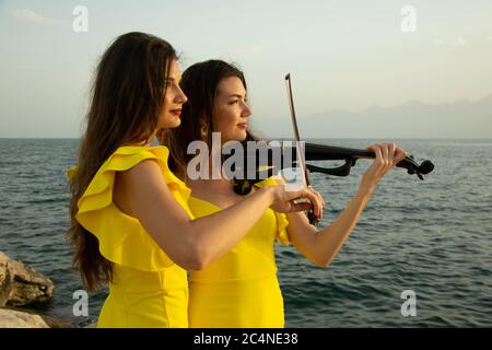 Two beautiful girls violinists in yellow concert dresses are playing electric violins with sunset, mountains and Mediterranean sea on the background. Stock Photo