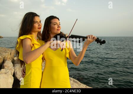 Two beautiful girls violinists in yellow concert dresses are playing electric violins with sunset, mountains and Mediterranean sea on the background. Stock Photo