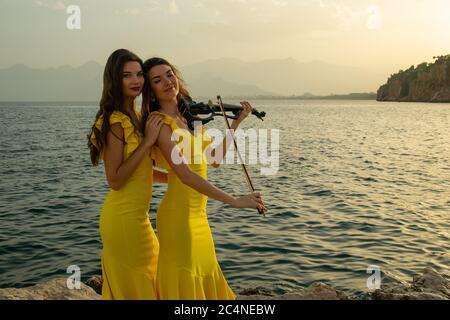 Two beautiful girls violinists in yellow concert dresses are playing electric violins with sunset, mountains and Mediterranean sea on the background. Stock Photo