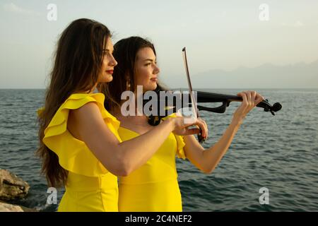 Two beautiful girls violinists in yellow concert dresses are playing electric violins with sunset, mountains and Mediterranean sea on the background. Stock Photo