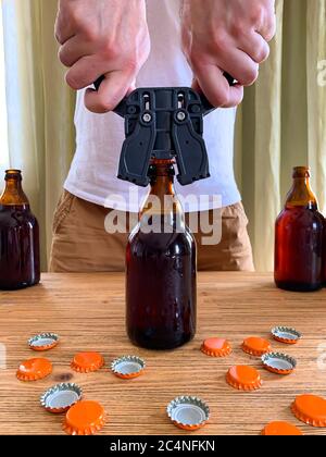 Craft beer brewing at home, man closes brown glass beer bottles with plastic capper on wooden table with orange crown caps. Vertical image Stock Photo