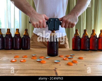 Craft beer brewing at home, man closes brown glass beer bottles with plastic capper on wooden table with orange crown caps. Horizontal image Stock Photo