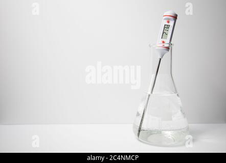 A digital thermometer measuring the temperature of a liquid in a glass erlenmeyer flask on a white background. Science experiment. Stock Photo
