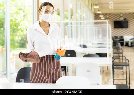 Portrait attractive asian waitress wear face mask and face shield hold alcohol and wet wipe for cleaning table before welcome customer. New normal hyg Stock Photo