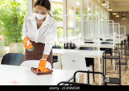 Portrait attractive asian waitress wear face mask and face shield cleaning table with alcohol and wet wipe before welcome customer. New normal hygiene Stock Photo