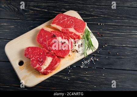 Three pieces of juicy raw beef with rosemary on a cutting board on a black wooden table background. Meat for barbecue or grill sprinkled with pepper and salt seasoning Stock Photo
