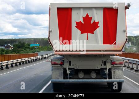 Big dirty truck with the national flag of Canada moving on the highway, against the background of the village and forest landscape. Concept of export- Stock Photo