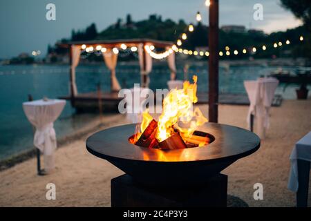 Grill with flames inside. Round table-cooking surface. On the beach, in the background of the gazebo by the water with garlands, in the twilight light Stock Photo