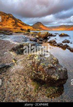 Rocks On Shoreline Of Wastwater Lake With Beautiful Golden Light On Mountains And Dark Dramatic Clouds. Lake District National Park, UK. Stock Photo