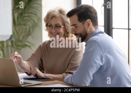 Smiling mature businesswoman mentor teaching intern, pointing at laptop screen Stock Photo