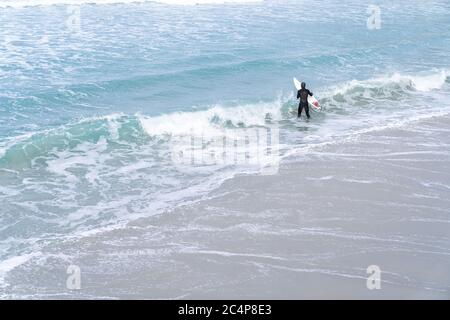 Surfer wearing wetsuit with surfboard watching ocean waves crash over rocks at St. Clair beach, Dunedin, New Zealand. Stock Photo