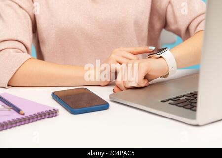 Closeup of female employee looking at smart watch on her hand while working on laptop in office, checking hour to take break, touching screen to turn Stock Photo