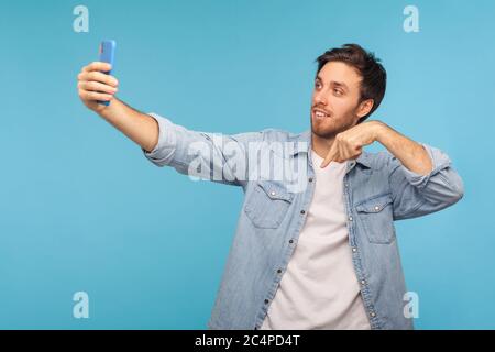 Portrait of handsome blogger man in denim shirt talking on video call and pointing down to subscribe, having online conversation on mobile phone, taki Stock Photo