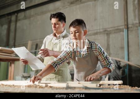 Middle-aged father in apron reading blueprint to son who assembling wooden furniture in workshop Stock Photo