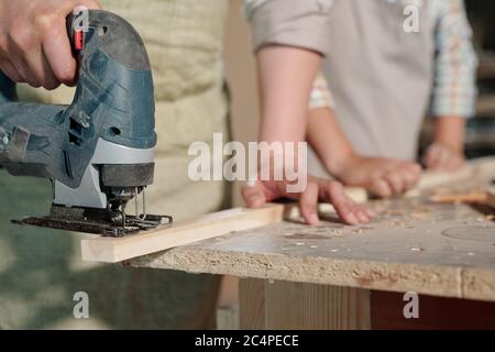Close-up of unrecognizable carpenter holding plank and using jigsaw while cutting it in workshop Stock Photo