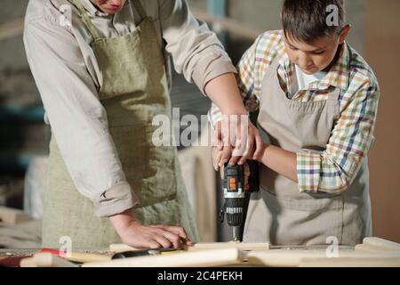 Close-up of skilled carpenter in apron pushing hands of son while helping him to drill wooden surface in workshop Stock Photo