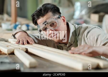 Concentrated middle-aged male carpenter in safety goggles examining surface of wooden planks in workshop Stock Photo
