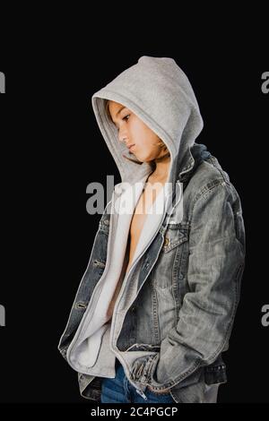 Eleven years old boy with hooded sweater and a jean jacket standing against a black background Stock Photo