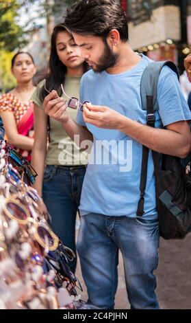 Indian young man choosing and buying sunglasses from outdoor street market with family at day time. Shoot location Sarojini Nagar, delhi, India.Indian Stock Photo