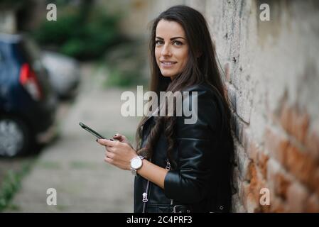 Young woman is smiling and holding phone by the brickwall. Stock Photo