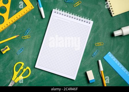 Blank squared notebook and school stationery on green chalkboard background. Flat lay, top view, overhead. Back to school, education concept. Stock Photo