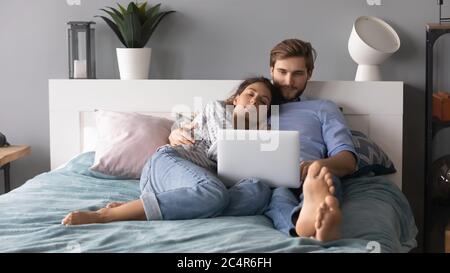 Young man and woman hugging, relaxing in bed, using laptop Stock Photo