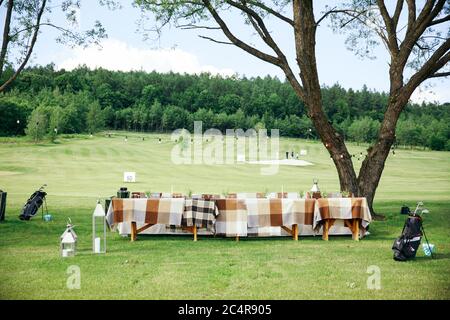 Long wooden table covered with white tablecloth, served with plates and glasses on green golf course. Beautiful serving for celebration outdoors. Stock Photo