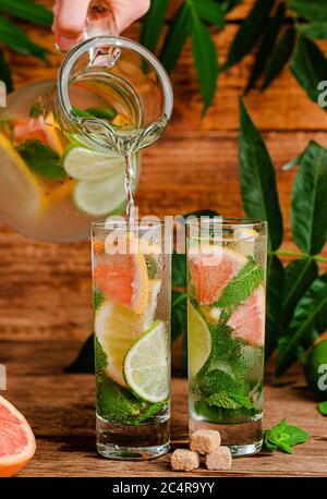 Iced summer drink with citrus fruits and mint pouring from transparent jug into glass on wooden background