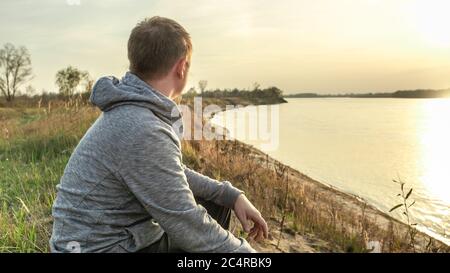 the guy is sitting on the riverbank and is looking thoughtfully at the setting sun. Evening sunset by the river Stock Photo