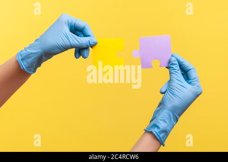 Profile side view closeup of human hand in blue surgical gloves holding yellow and purple puzzle pieses. indoor, studio shot, isolated on yellow backg Stock Photo