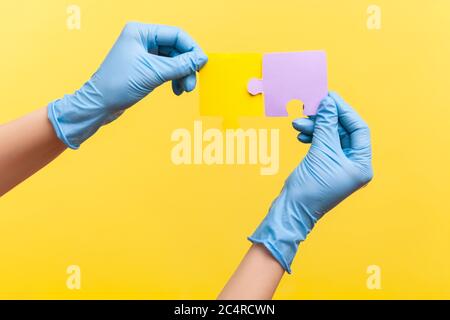 Profile side view closeup of human hand in blue surgical gloves holding yellow and purple puzzle pieses. indoor, studio shot, isolated on yellow backg Stock Photo