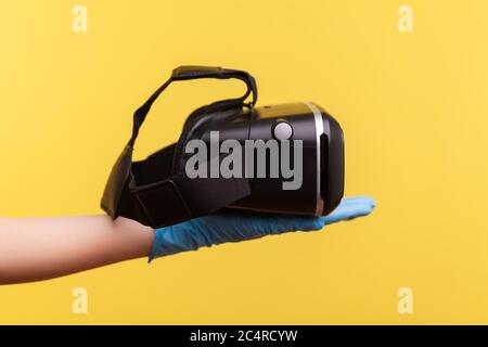 Profile side view closeup of human hand in blue surgical gloves holding virtual reality headset. indoor, studio shot, isolated on yellow background. Stock Photo