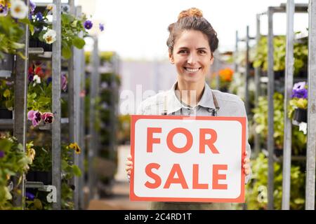 Waist up portrait of smiling female gardener holding FOR SALE sign while standing in greenhouse at flower plantation, copy space Stock Photo
