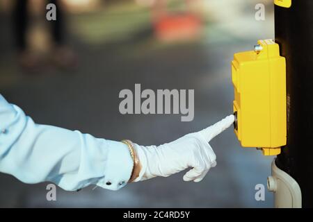 Life during coronavirus pandemic. Closeup on female in blue blouse with gloves pressing the traffic light button to cross the street outdoors in the c Stock Photo