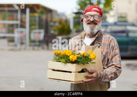 Waist up portrait of cheerful senior gardener holding flowers and looking at camera while standing by tree market outdoors, copy space Stock Photo