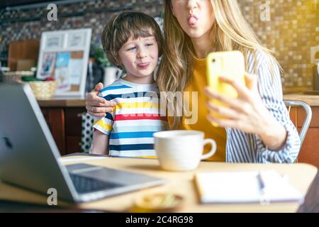 Mother and son show tongue, make selfie looking at smart phone. Having fun together at home. Woman working from home and online education with kid wit Stock Photo