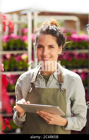 Vertical waist up portrait of smiling young woman holding digital tablet and looking at camera while enjoying work in plantation outdoors, copy space Stock Photo