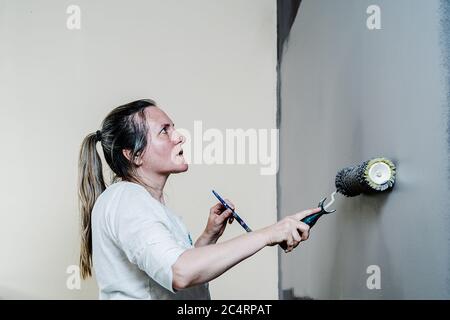 Woman catching with a hand a paint roller full of grey painting and with the other a brush. She is upping and downing the roller covering the wall wit Stock Photo