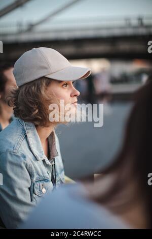 Beautiful blonde girl in a cap with short hair sits in a group of girls and looking away. Loneliness among friends. Selective focus. Stock Photo