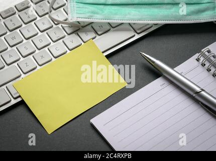 Office desk table with computer keyboard, notepad, pen, blank yellow paper and medical face mask. Top view or flat lay. Stock Photo