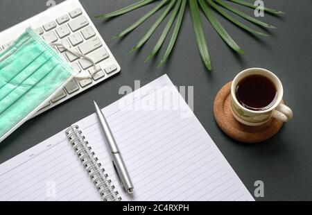 Top view of office desk with computer keyboard, medical face mask, notepad, coffee and green plant. Stock Photo